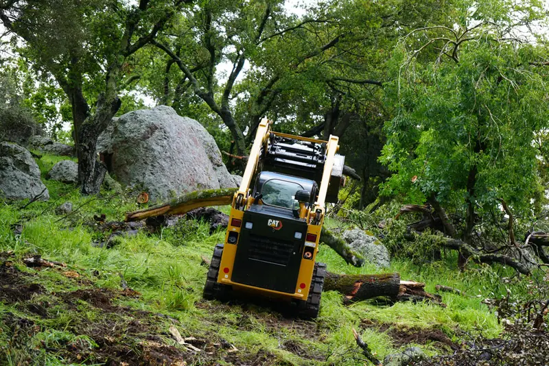 Tracked loader working through fallen oak limbs on a wooded, rocky hillside cleanup